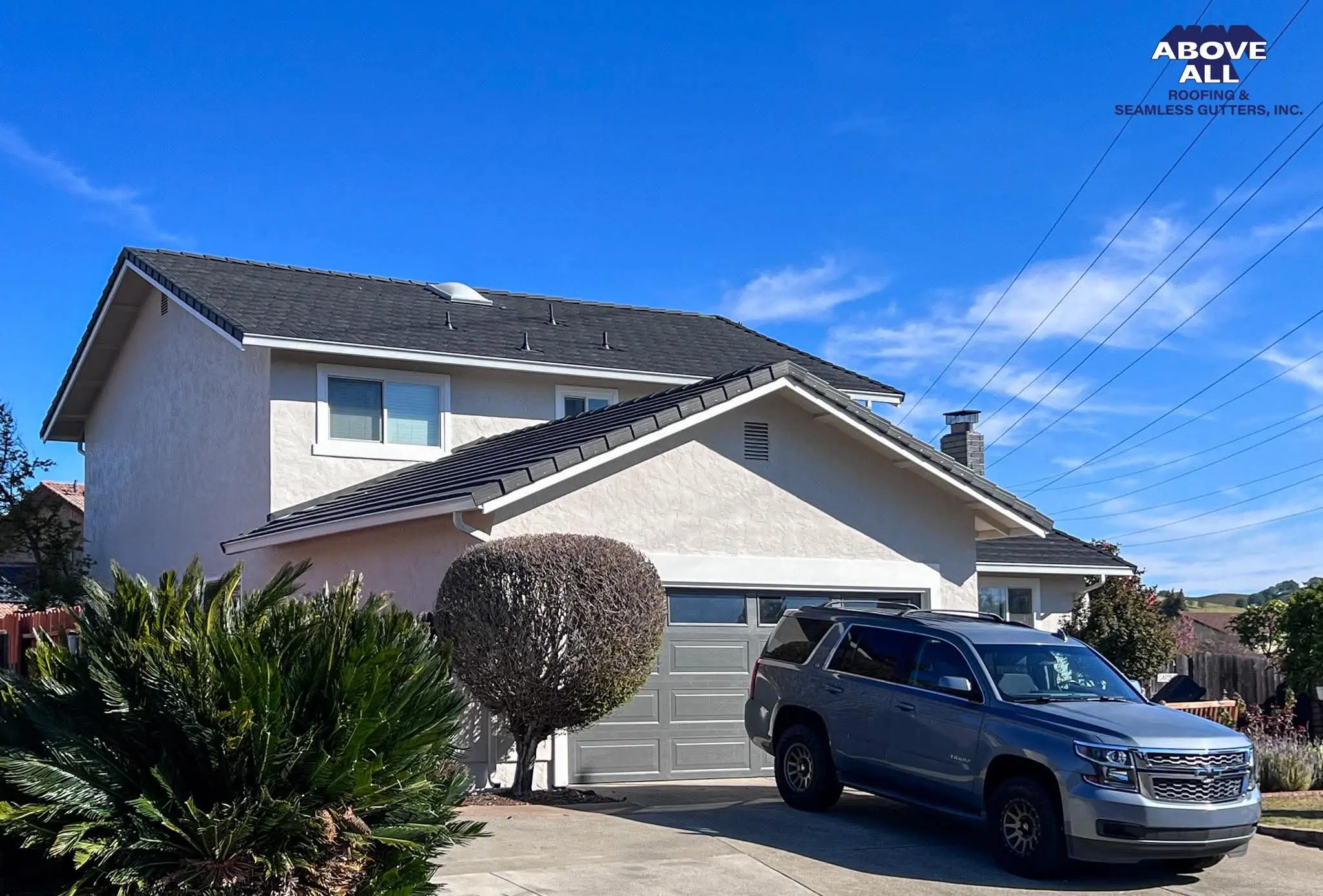 19112 A two-story beige house with a gray roof and garage, a neatly trimmed bush in front, and a silver SUV parked in the driveway under a blue sky. The Above All Roofing & Seamless Gutters, Inc. logo is in the top right corner.